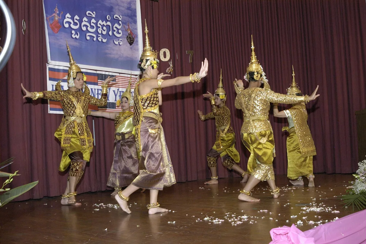 Dancers in gold and green classical costumes and tall gold crowns, mid-movement on a polished stage in front of a maroon curtain.