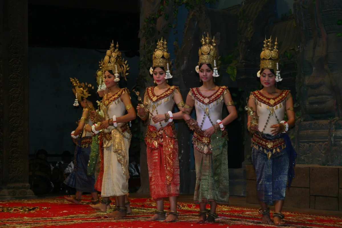 Five apsara dancers in a line, in gold crowns and cream bodices, with skirt-cloths in red, green and blue on a red carpet.