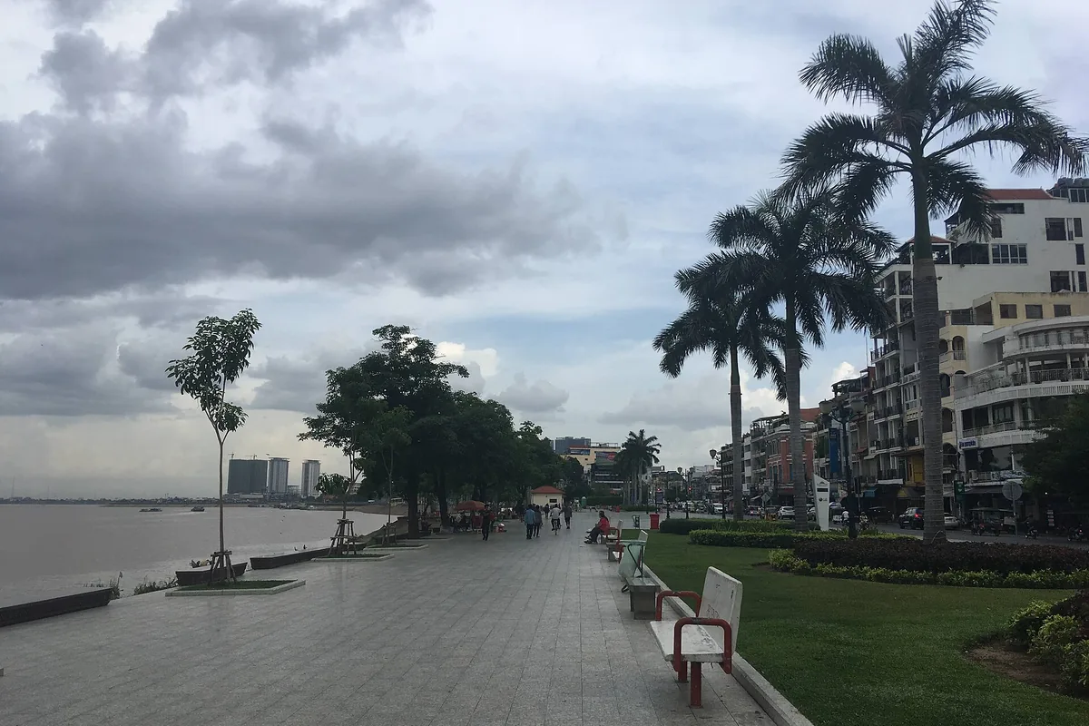 The paved promenade beside the Tonlé Sap in Phnom Penh under a cloudy sky, with royal palms and buildings on the right and the river on the left.