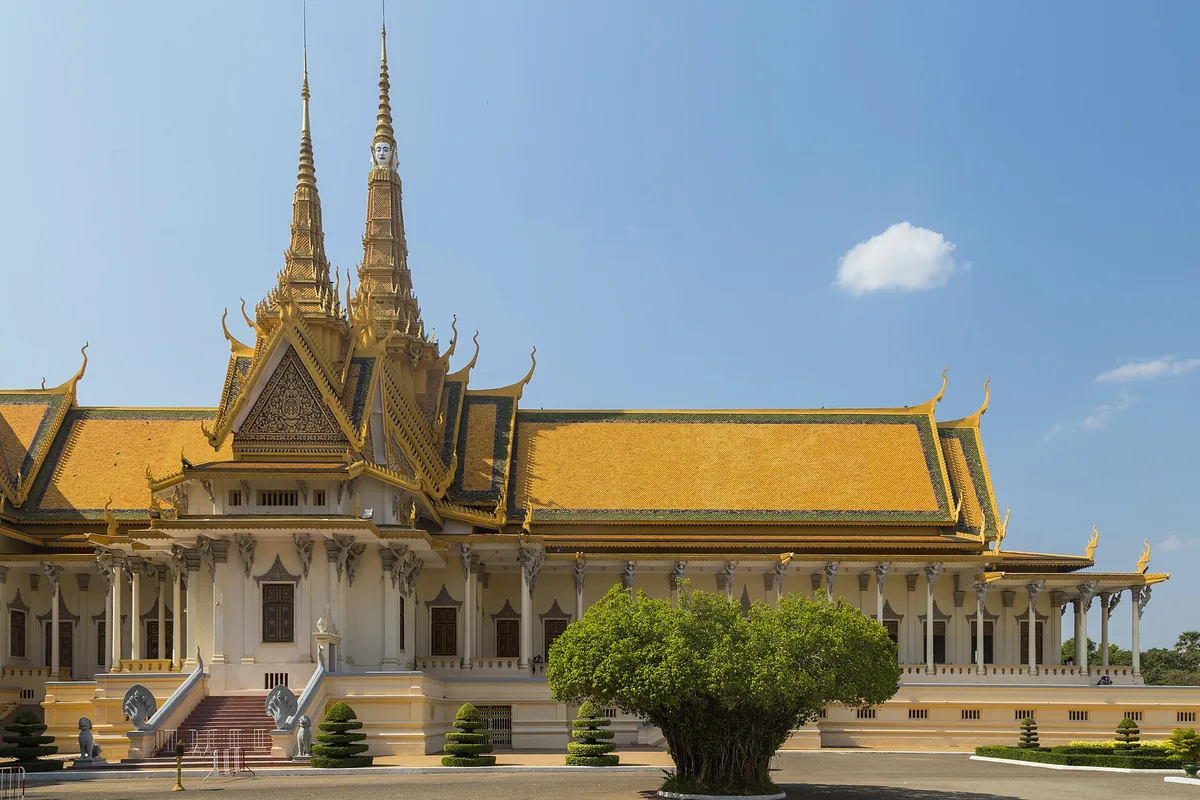 The front of the Throne Hall of the Royal Palace in Phnom Penh, a cross-shaped building with golden roofs and twin spires, a clipped tree in the foreground.